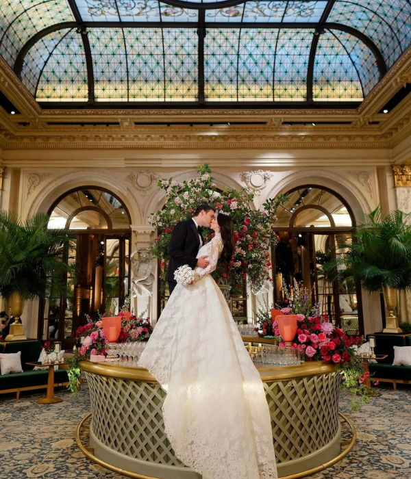 Bride and groom share a romantic kiss atop a gold table decorated with flowers inside an elegant ballroom surrounded by lush greenery and ornate architecture.