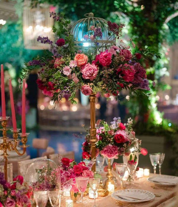 Luxurious wedding reception table decorated with vibrant pink and red floral centerpieces, glowing candles, crystal glassware, and an ornate birdcage arrangement under ambient evening lighting.