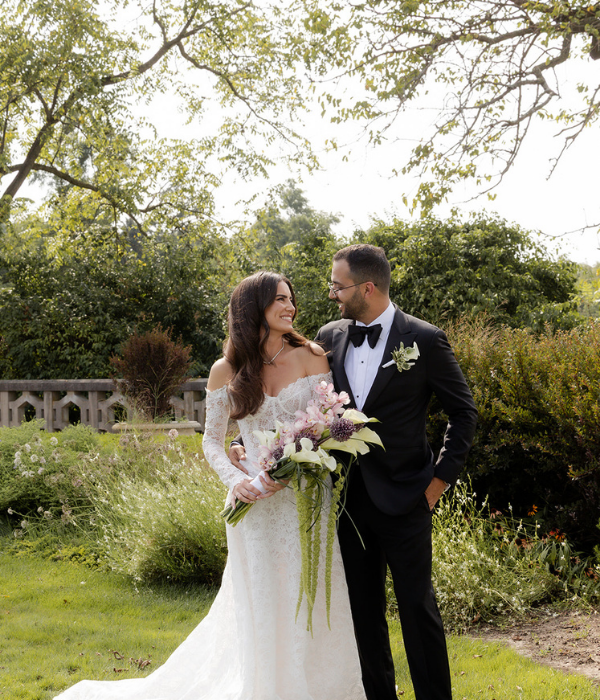 Bride and groom standing in a sunlit garden, smiling at each other while holding a pastel bouquet, surrounded by lush greenery and soft natural light.