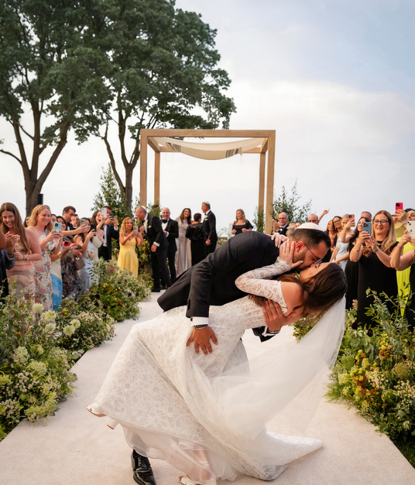 Groom dips and kisses bride at the end of a joyful outdoor wedding ceremony surrounded by cheering guests capturing the moment on their phones.