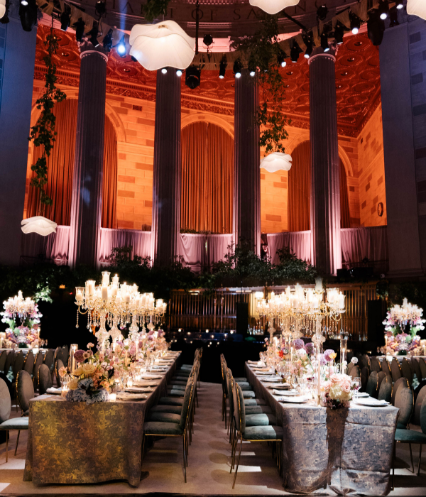 A dramatic reception space with towering columns, warm uplighting, long banquet tables, and ornate crystal candelabras surrounded by floral centerpieces.