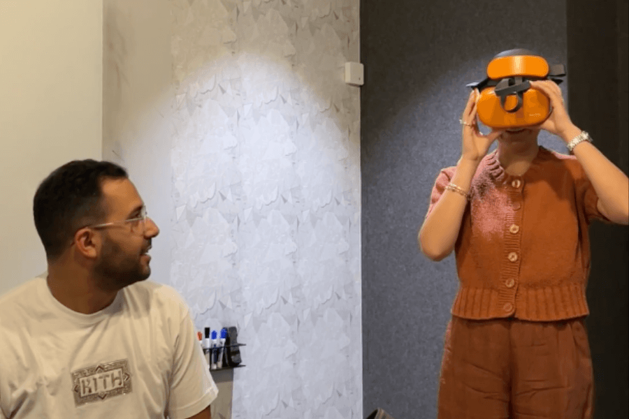 A woman testing a bright orange Reloom VR headset while a man watches, showcasing an immersive virtual reality demo and memory-capture experience.